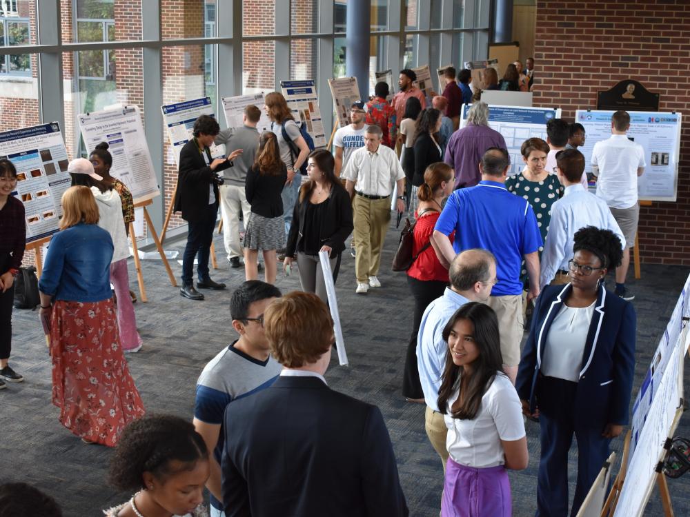 Scientific posters on easels line a large room, with windows on one side. People are grouped together in two or threes, discussing the research on the posters. 