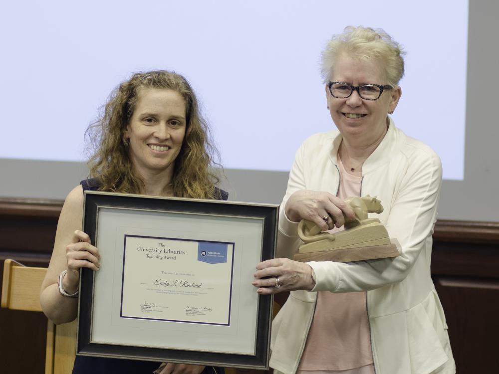 two women standing, one holding award certificate, other presenting small sculpture of Nittany Lion as award