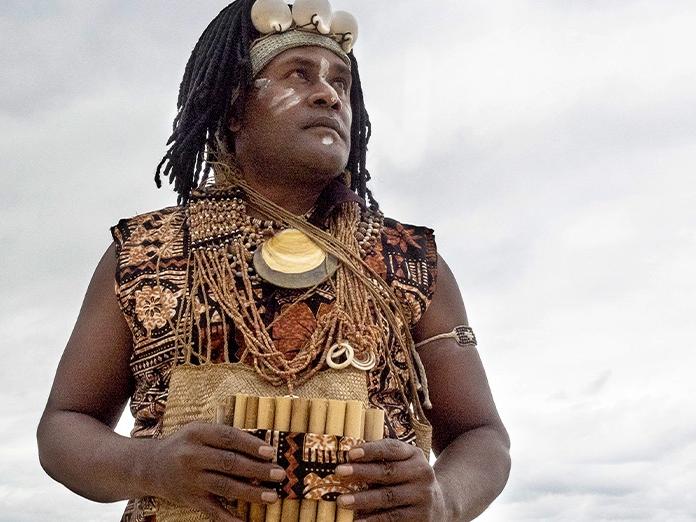 A man wearing shells and beads on his bare chest holds a pan pipe.