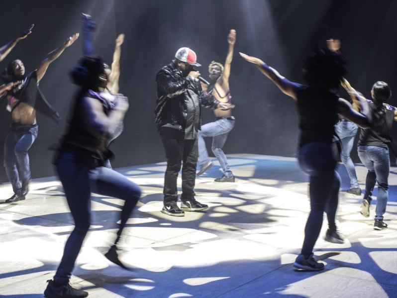 A group of dancers move in a circle around a man singing into a microphone.