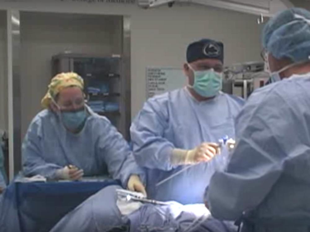 Dr. Gerald Harkins holds an operating tool and looks at an assistant during a procedure featured on Surgery Live! He wears a surgical gown, gloves, cap and mask. A nurse in a gown and mask next to him readies surgical instruments on a tray.
