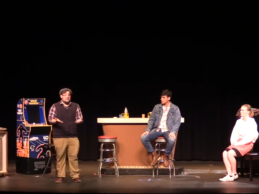 two men are sitting at a bar, one man talking animatedly, and a woman is sitting backward at a piano bench listening