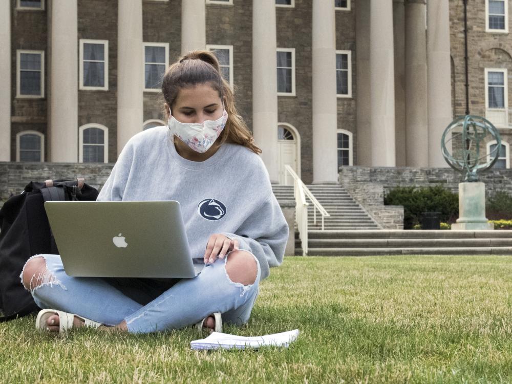 A female student sits in front on the Old Main lawn wearing a face mask.