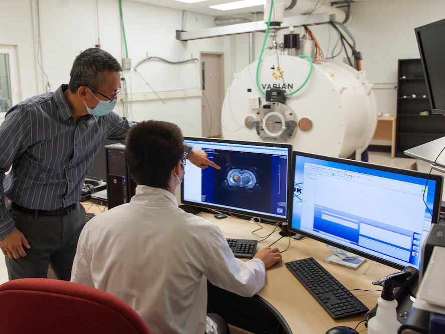 One man, standing, points to an image on a computer screen to a man, who is sitting at a desk. A large white tube is shown behind them. 
