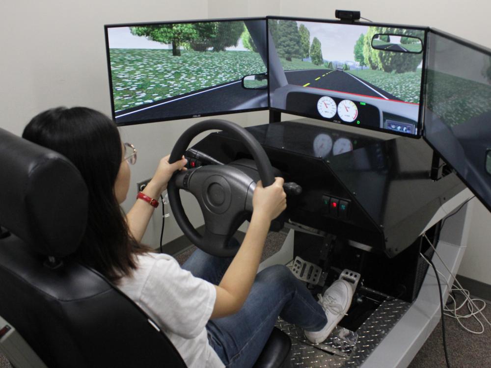 A student demonstrates a driving simulator that consists of a car seat, a wheel, and three screens showing a road on them. 