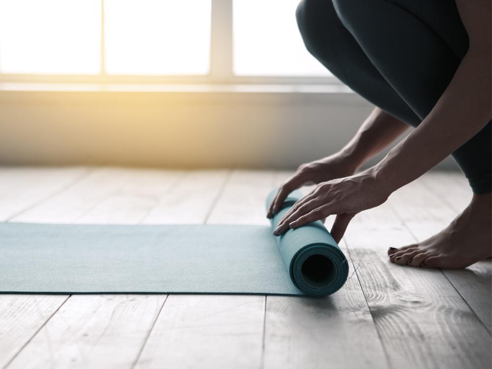 A close-up of a woman’s hands as they roll up a yoga mat, which is on a hardwood floor. Windows are in the background.