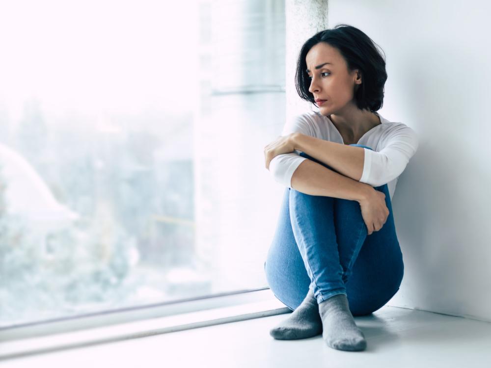 A woman sits on the floor, her arms resting on her legs. She looks out the window on a sunny day.