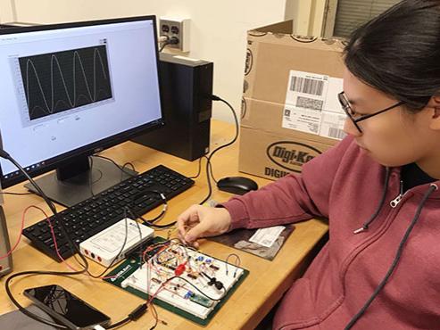 A student in a zip-up sweatshirt and glasses works on a circuit board in front of a computer screen displaying a graph 