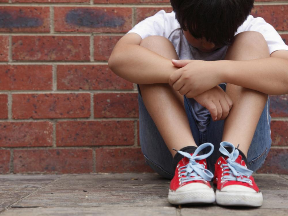 A young boy sits on a sidewalk, leaning against a brick wall, looking down at the ground.