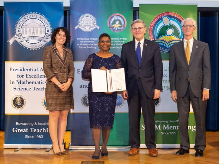 Two women and men stand on a stage, smiling at the camera. The African-American woman holds an award. 