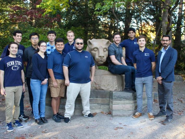 A group of graduate students and a professor stand next to the Nittany Lion statue.