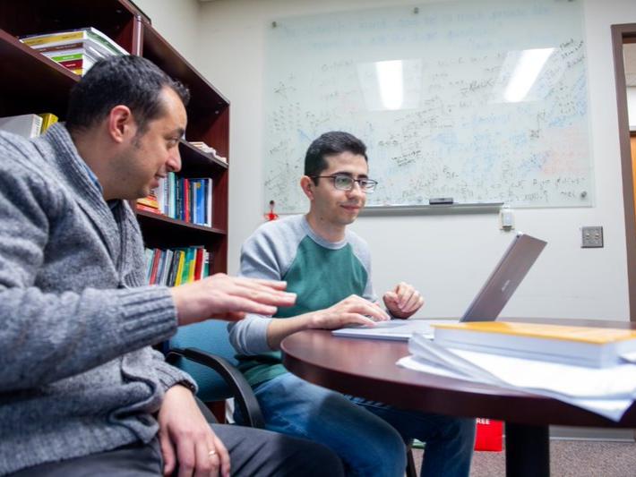 Man in a gray sweater speaks to man in green shirt while looking at laptop.