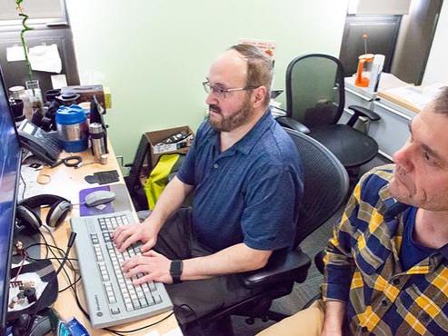 A man in a blue shirt with glasses and a man in a plaid shirt to his right work at a computer