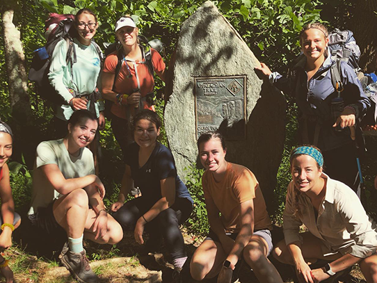 Nine women dressed in hiking gear pose for a photo near a rock. 