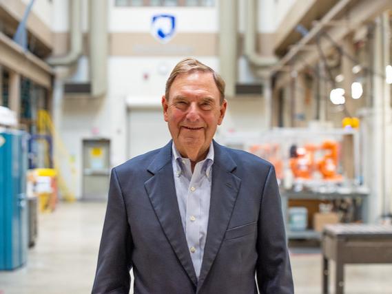Man stands in the FAME Lab, smiling for the camera while wearing a blue shirt and a suit jacket with the Nittany Lion shield blurred in the background.