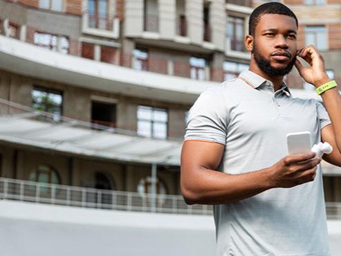 A man wearing a polo shirt and a fitness tracker on his wrist holds a smartphone while adjusting his headphones. 