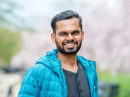 A man wears a light blue jacket and smiles at the camera in front of a blurred background of cherry blossom trees 