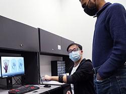 A person in a black zip-up sweatshirt and a face mask models a piece of technology on his wrist in front of a computer while a person in a blue shirt and a face mask looks on.