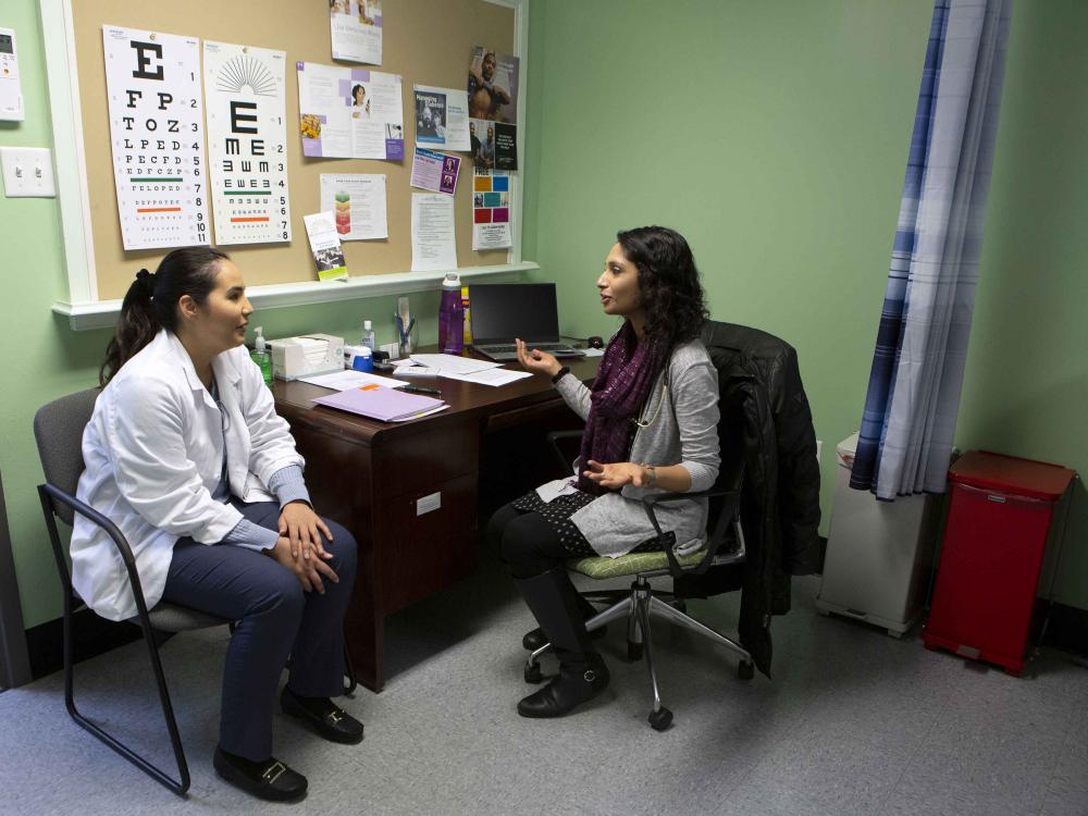 Dr. Leesha Helm, seated at a desk, gestures toward Penn State College of Medicine third-year student Maria Escudero during a shadowing visit at Beacon Clinic. A bulletin board with an eye chart and other papers on it is on the wall behind the desk.