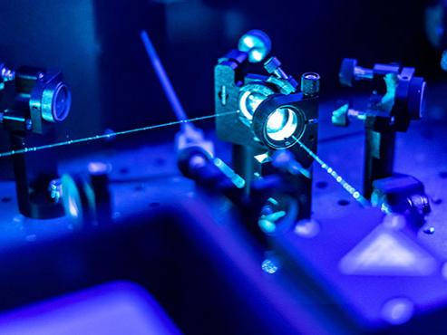 Beams of blue light shine out from a laser made of two optical lenses. Two similar lenses are on either side of the laser, and the entire image is tinted blue. 