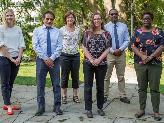 Four women and three men stand on an outside walkway with greenery in the background to pose for a photo 