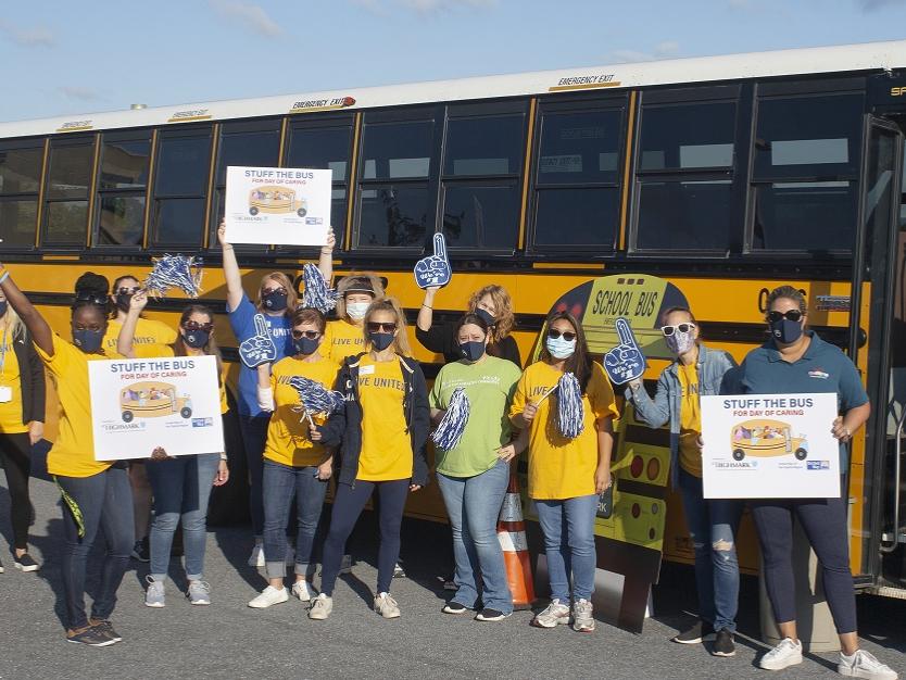 A group of 14 women stand in front of a school bus holding “Stuff the Bus” signs, pom-poms and foam fingers. Ten of them are wearing United Way T-shirts. Behind them is a Penn State Health pop-up tent.