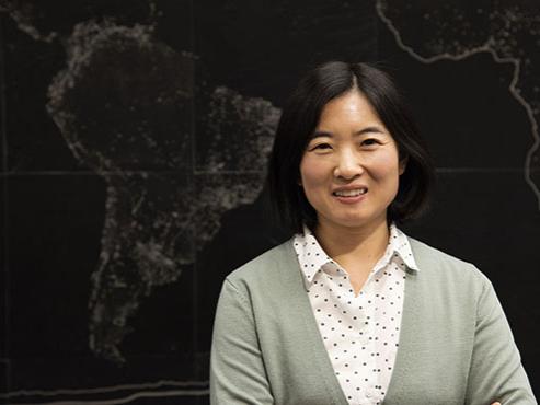 A woman in a polka dot shirt and green sweater smiles and poses with her arms crossed in front of a world map