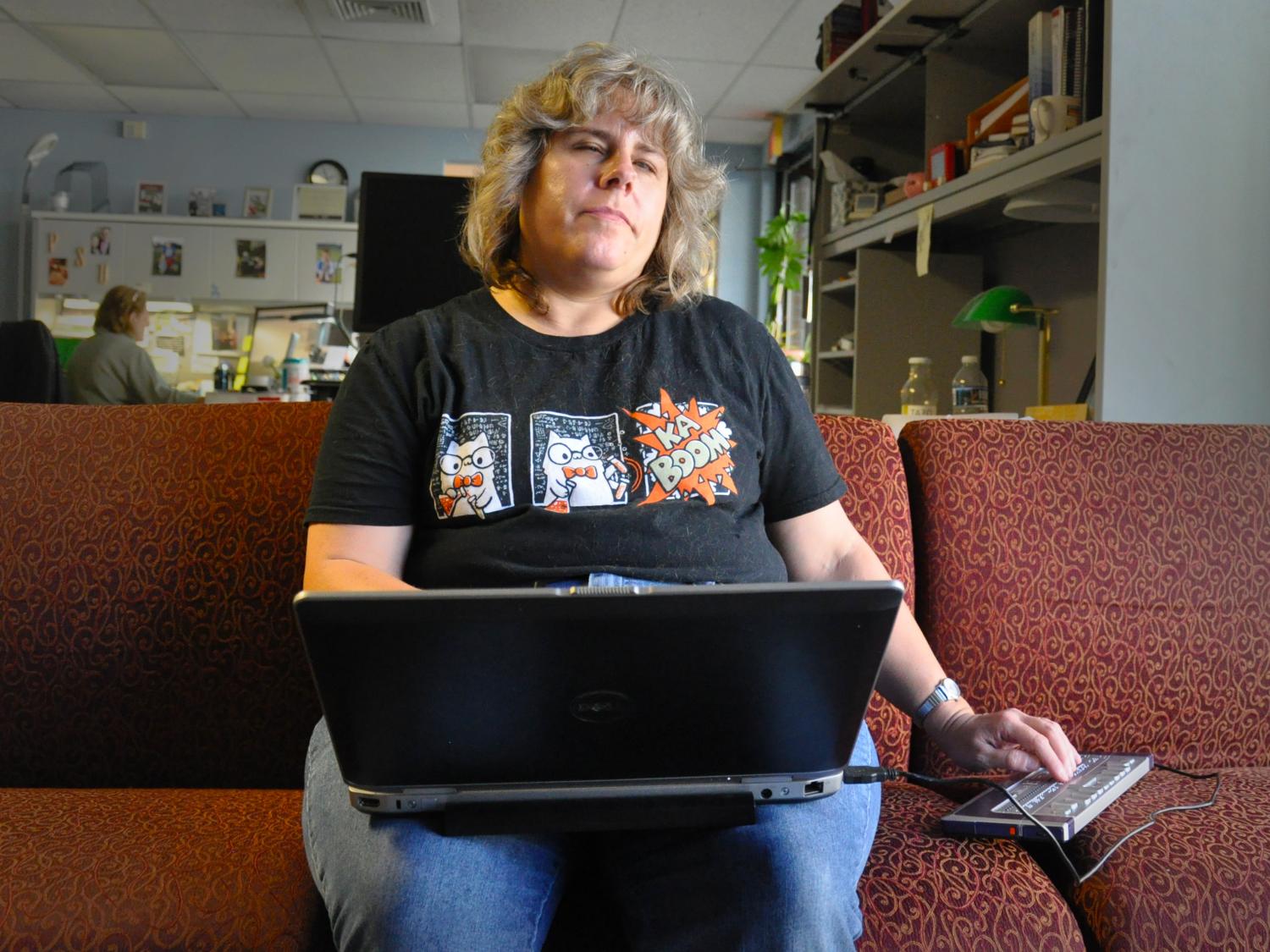 A woman sits on a couch with a laptop on her lap and a braille keyboard to her right