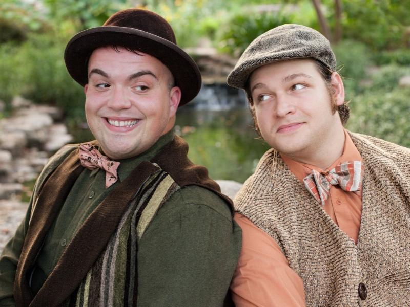 Two actors wearing hats smile slyly at each other and sit shoulder to shoulder.