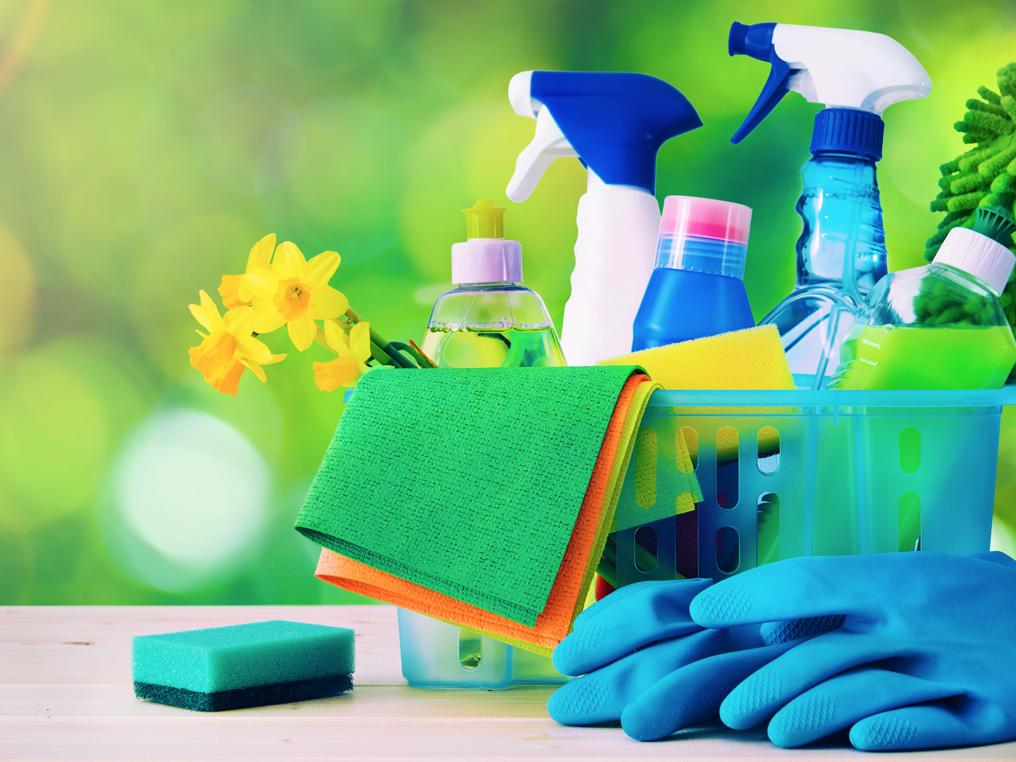 A plastic container with various cleaning supplies, including spray bottles and sponges, is on a table. Also sitting on the table is a pair of rubber gloves and a small sponge. Green foliage is in the background, out of focus.