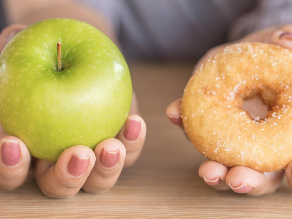 A close-up of two outstretched hands – one holding an apple tilted toward the camera, the other holding a doughnut tilted toward the camera.