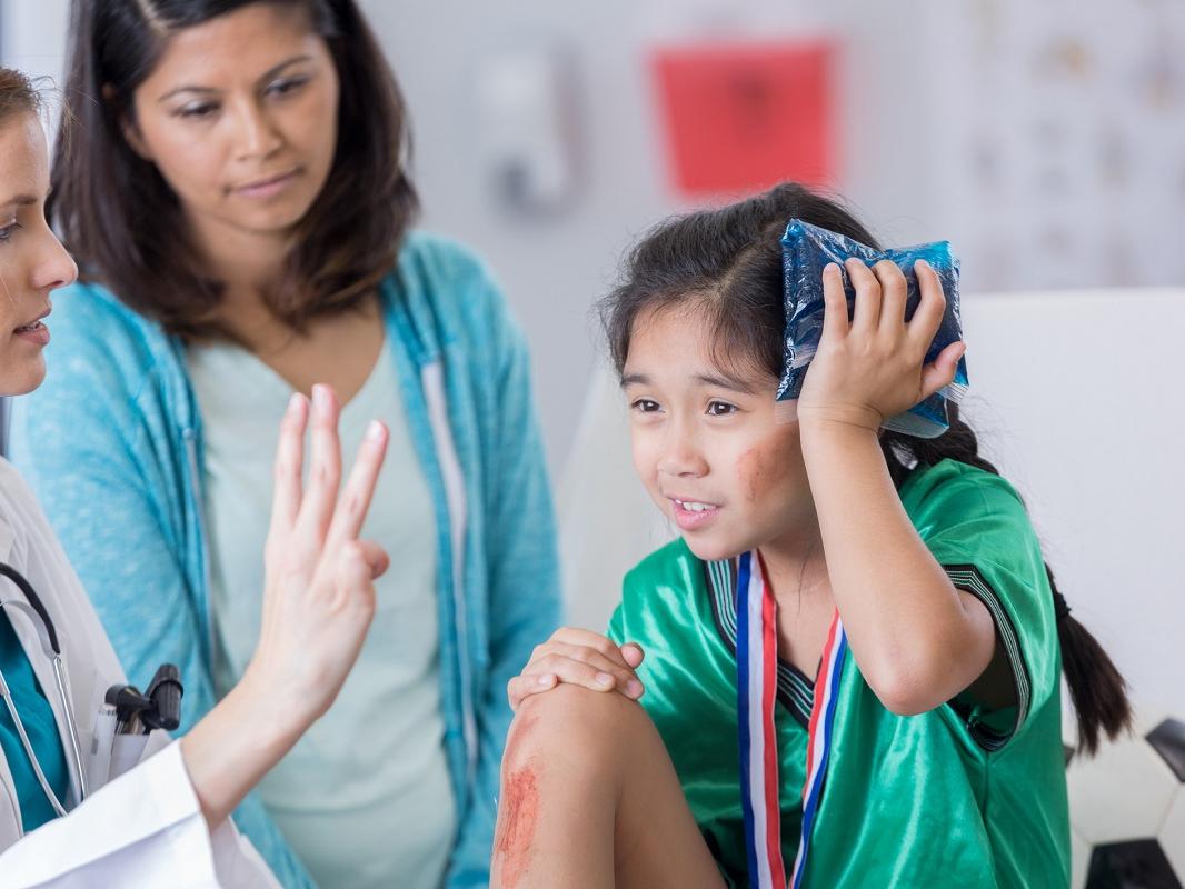 Serious female ER doctor holds up three fingers while talking with injured female elementary age soccer player. The doctor is asking the girl how many fingers she is holding up. The girl is wearing a medal and soccer uniform.