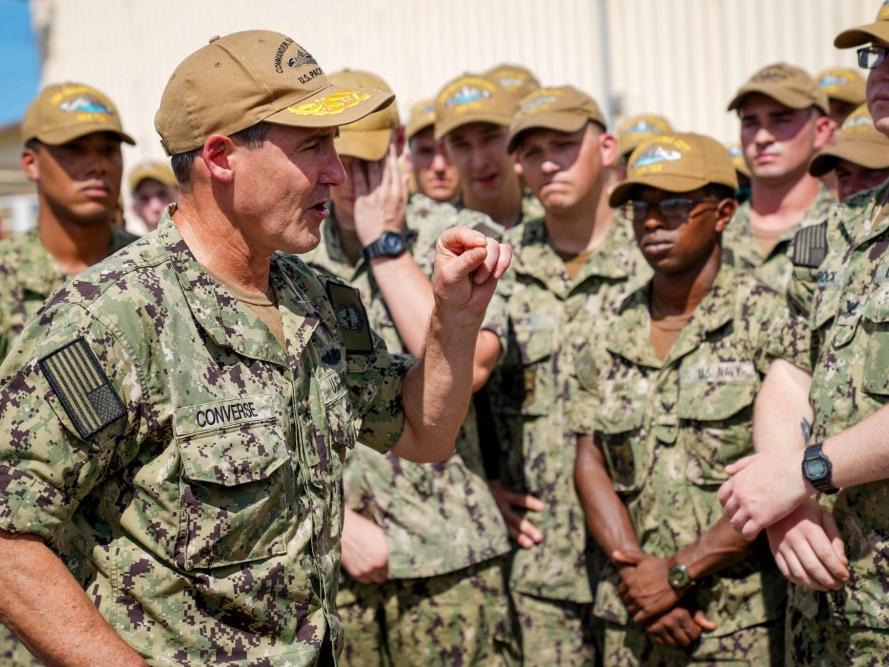 A military officer addresses troops with his hand raised, all dressed in camouflage.