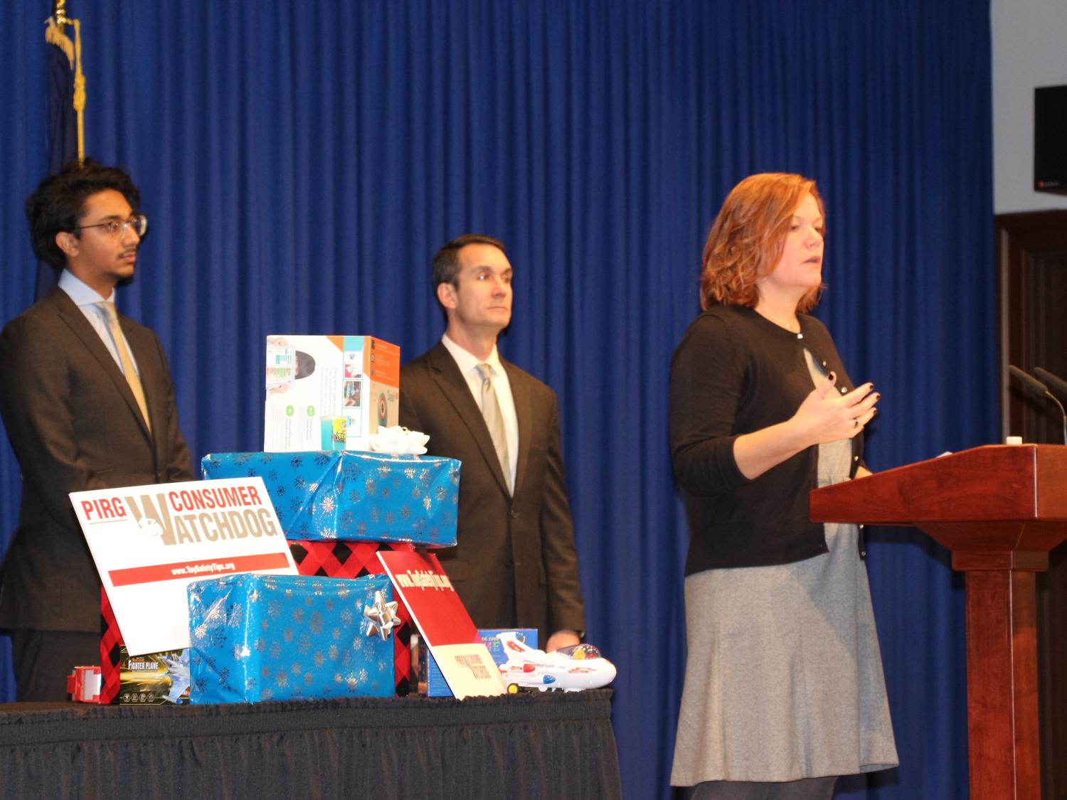 Pediatric Trauma and Injury Prevention manager Amy Bollinger, right, a woman with shoulder-length hair, stands at a podium discussing unsafe toys at a press conference. Behind her stand Reuben Mathew of PennPIRG, left, and Pennsylvania Auditor General Eugene DePasquale, right. Between the two men is a table with wrapped gifts.