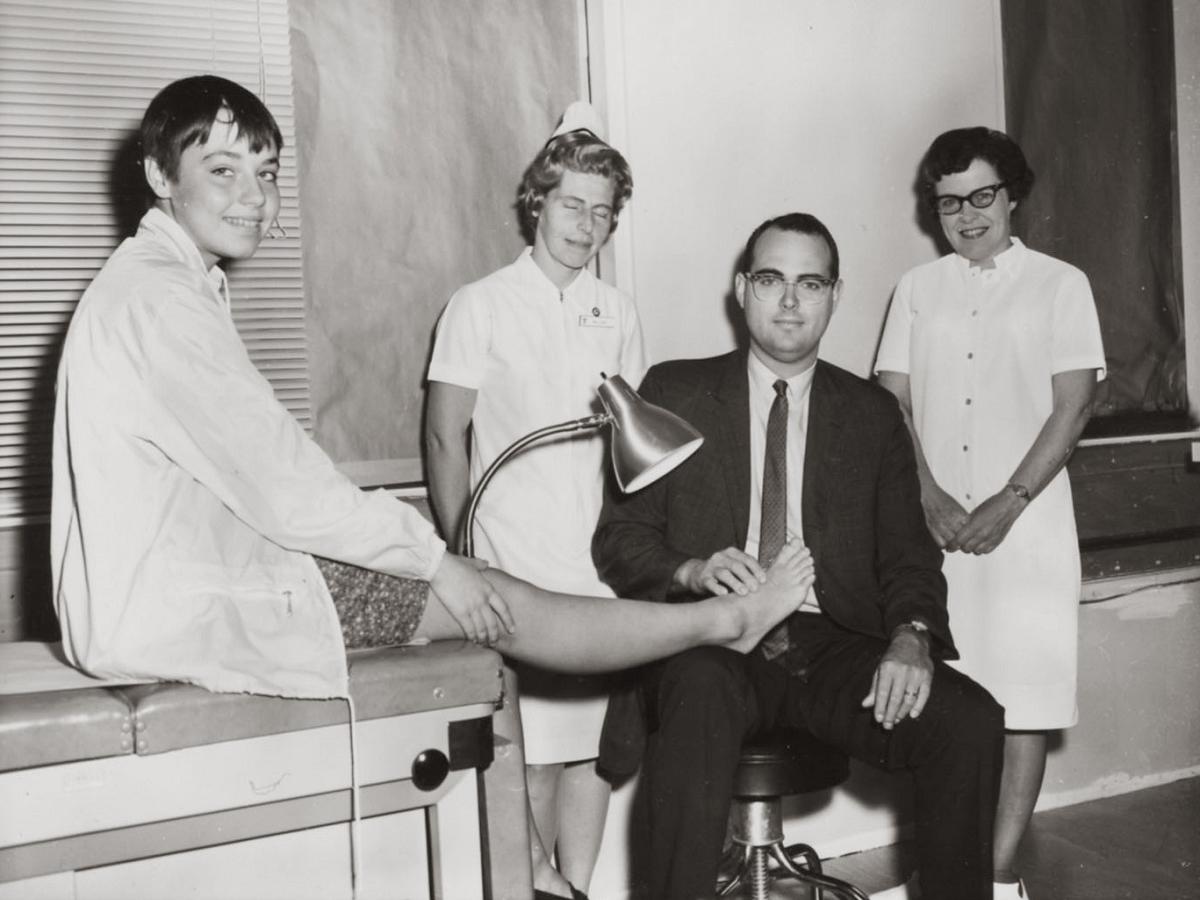 Dr. Willis W. Willard III, wearing a suit and tie, holds the foot of a young boy on an exam table. Two nurses, dressed in white dresses and nursing caps, stand in the background. The boy wears shorts and a jacket and is smiling.