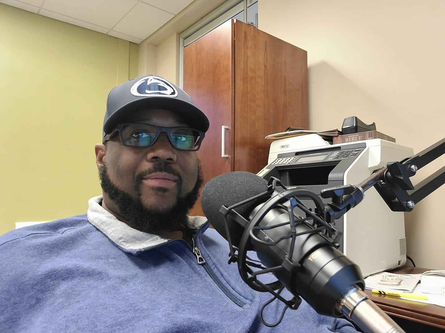 man sitting at microphone wearing a penn state hat. 