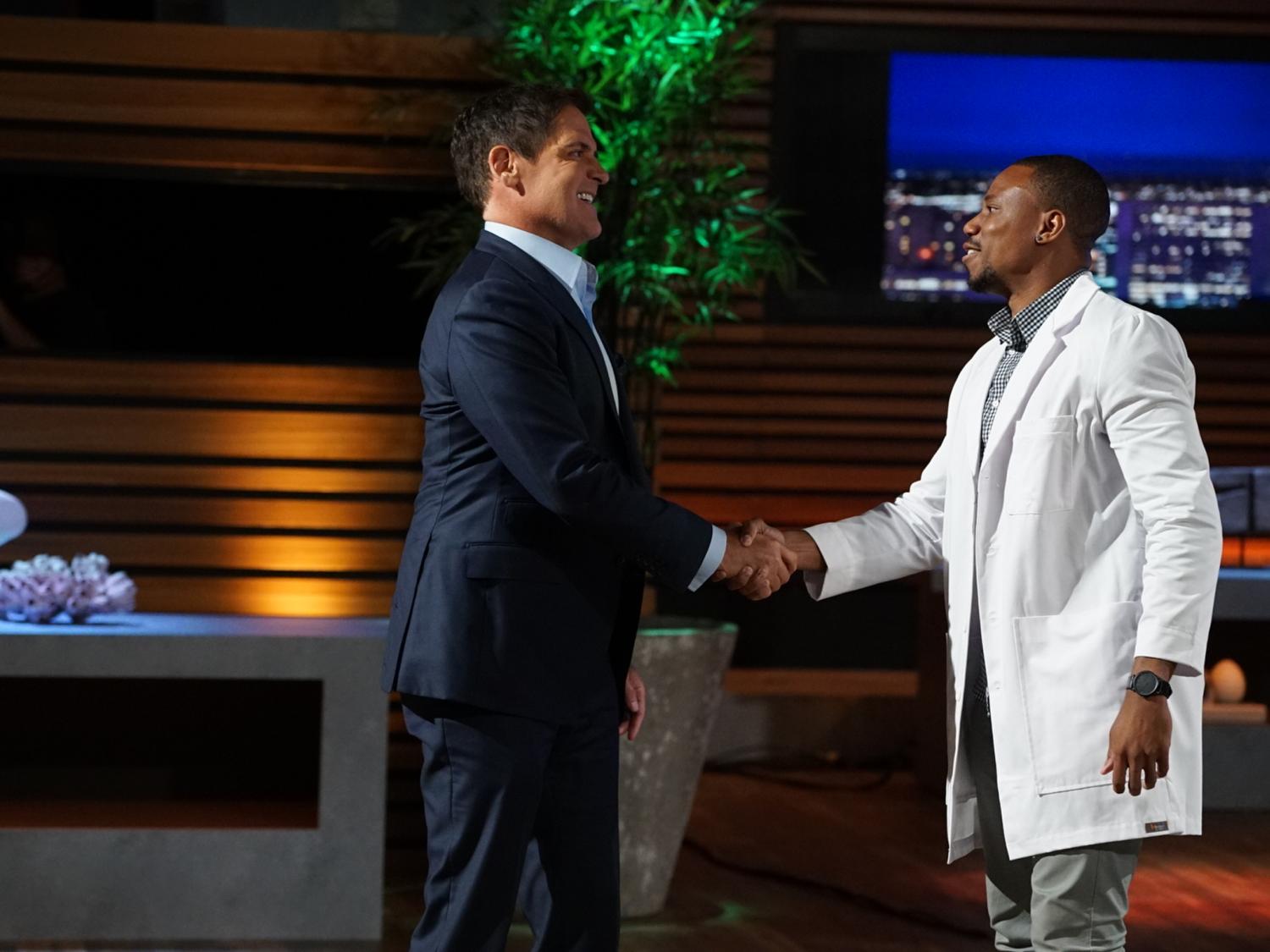 A man in a dark suit (at left) and a man in a white lab coat shake hands and smile. In the background is a TV set with wood paneling and a tall green plant.