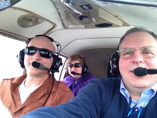 Three people are seated in an aircraft. Two men are nearest the camera. A woman is in the back seat. All three are wearing headsets.