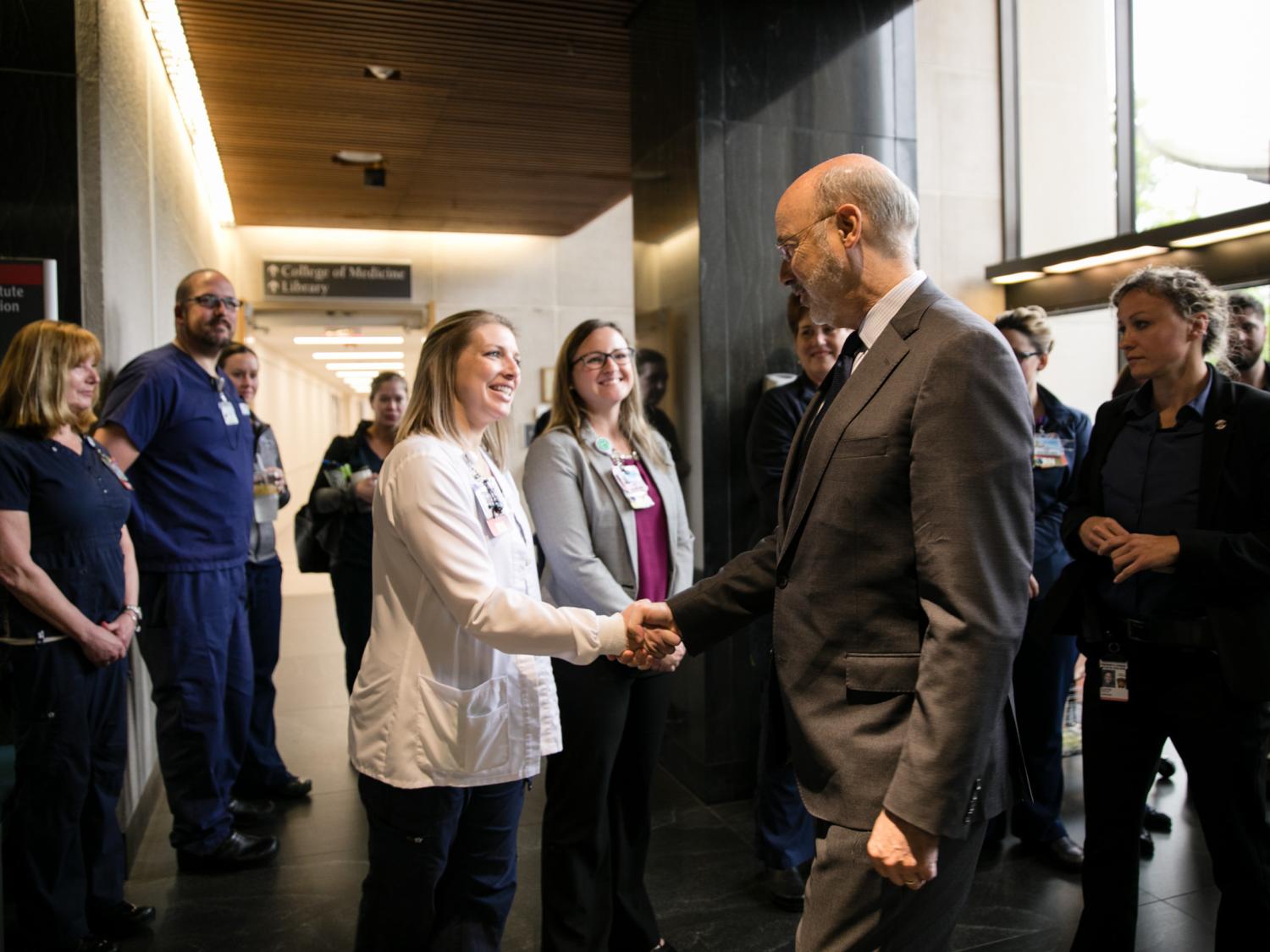 Gov. Tom Wolf, wearing a suit, shakes hands with a woman wearing a medical jacket. Other people in professional and medical attire look on. A hallway and large windows are in the background.