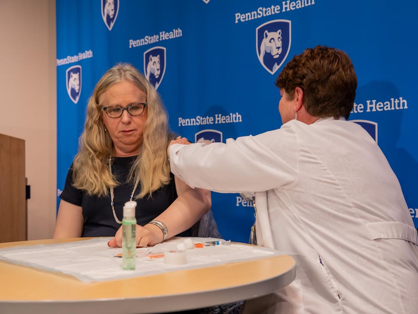 Two women sit at a table, one gives the other a flu vaccine in the upper arm.