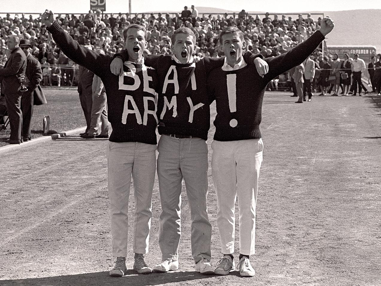 black-and-white photo of three young men cheering and standing on football field sidelines wearing sweaters that when combined spell out "Beat Army!"
