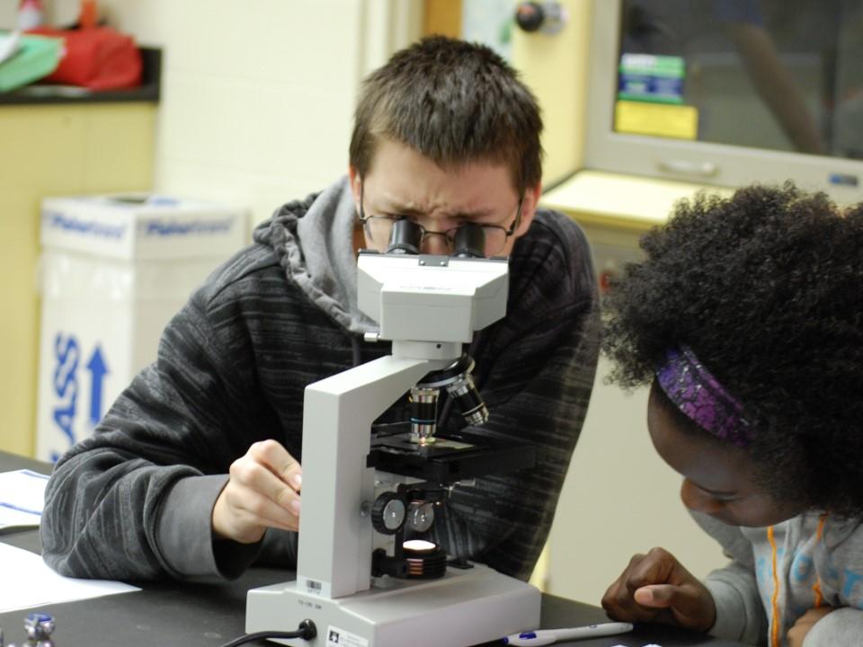 Two young students are pictured--one looking into a microscope and the other taking notes.