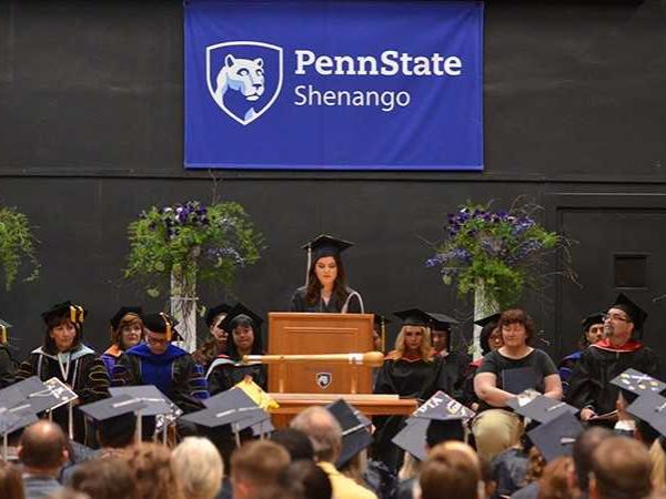 A student stands at a podium speaking while other faculty and staff sit on the stage watching.