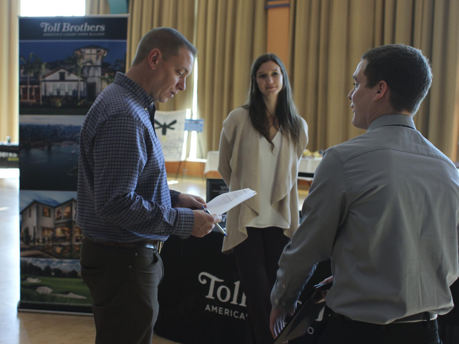A female student wearing a tan cardigan and a male student wearing a gray dress shirt both look at a man in a blue dress shirt, who is reviewing a resume.
