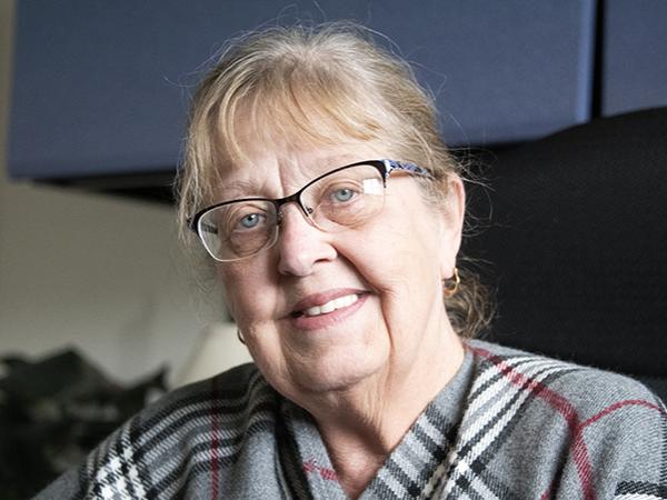 A woman, Mary Ann Walters, is seated at her desk at Penn State Fayette. She wears a striped shawl and glasses.