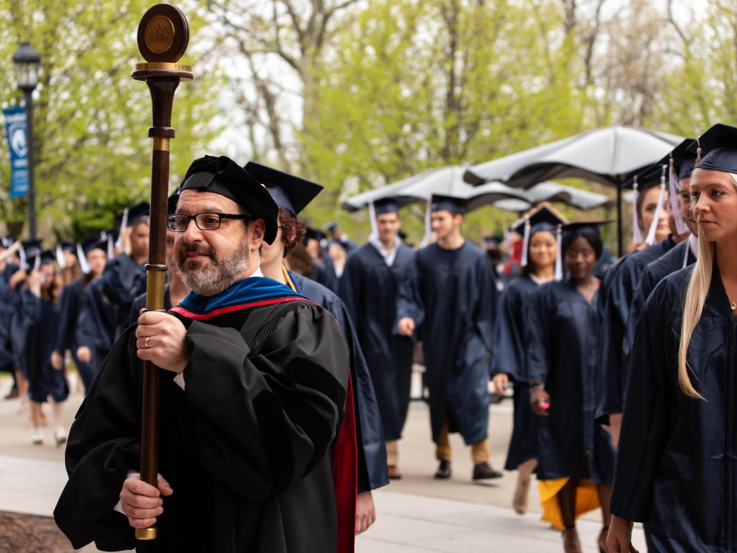 A male professor in a black academic robe carries a mace and leads two rows of graduates in dark blue caps and gowns to the graduation ceremony. 
