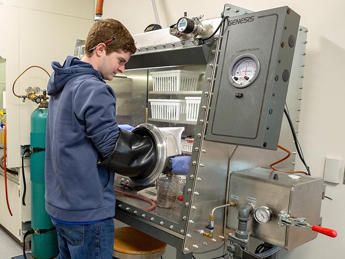 A student is working in front of a glass box. Rubber gloves, that are built into the box, extend up to his elbows as he reaches into the chamber.