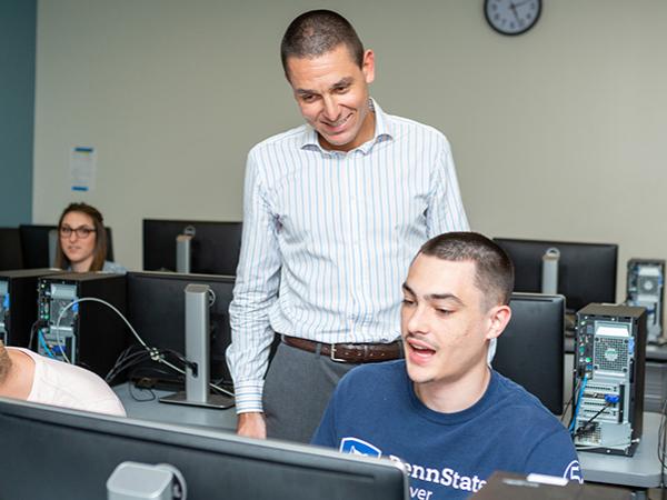 Dr. Andrea Patrucco, wearing a white shirt with blue vertical stripes, stands behind a student seated at a computer.