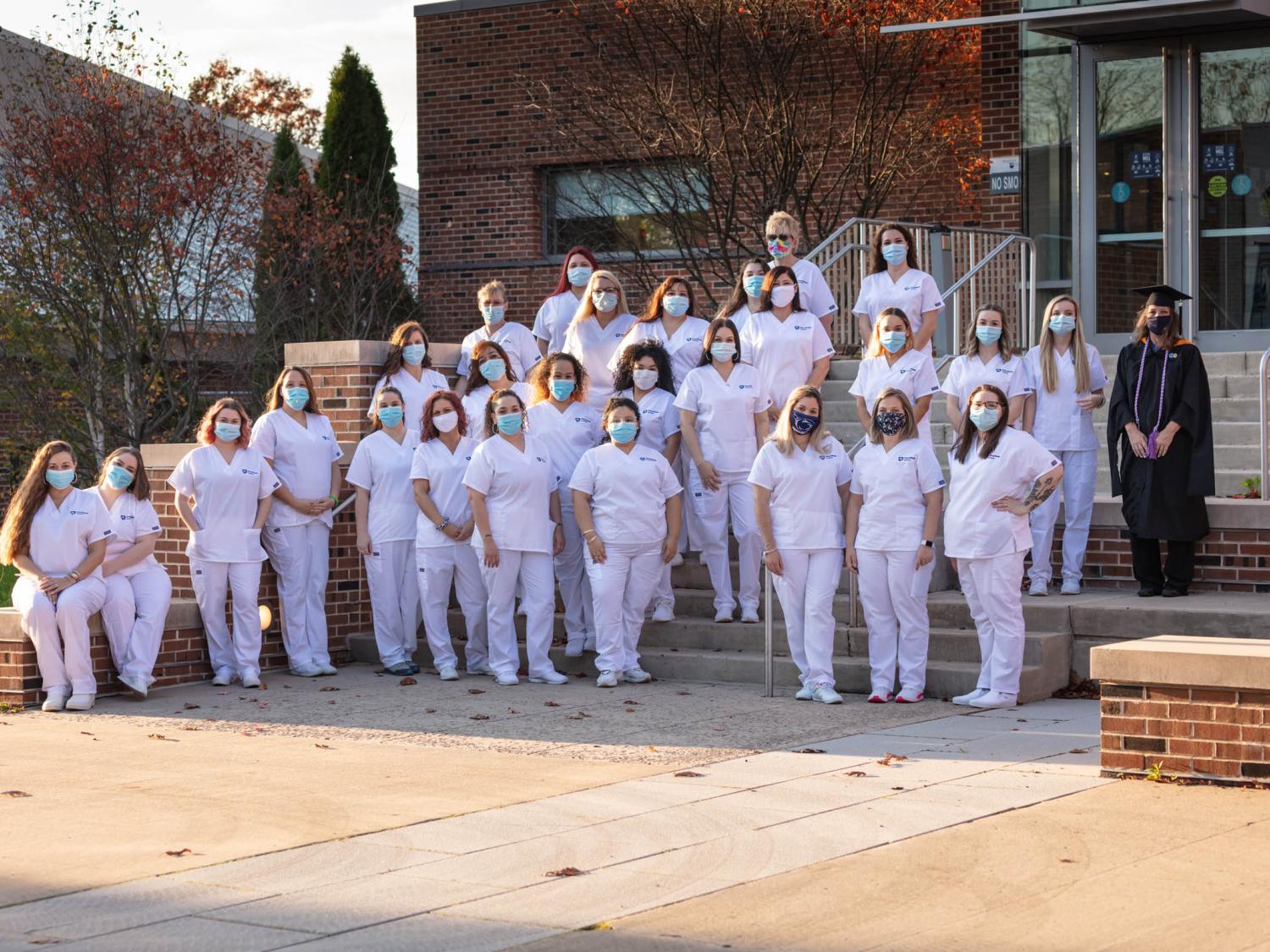 Rows of practical nursing students in white scrubs lined up on steps in front of campus building.