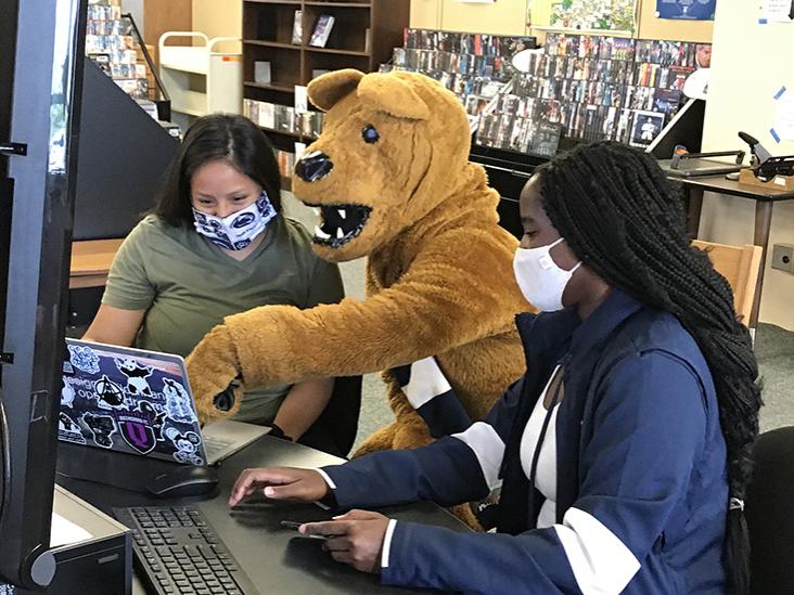 The Nittany Lion sits at a computer with two female students in the library.